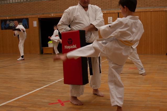 Junge in Karate-Duniform tritt gegen einen gepolsterten Schlagblock, während ein Trainer ihn anleitet.