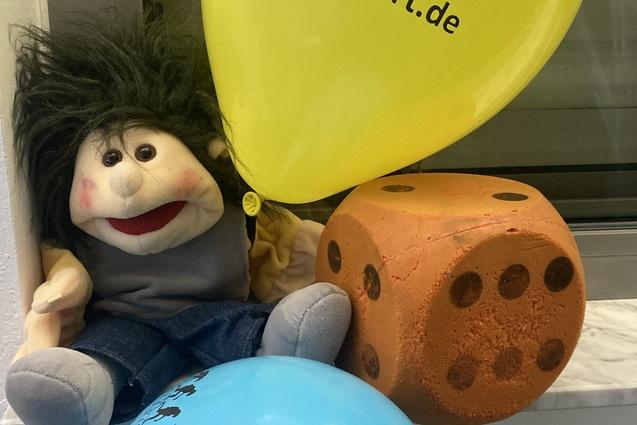 Plüschfigur mit frecher Frisur sitzt neben orangenem Würfel und blauen sowie gelben Luftballons auf einer Fensterbank.