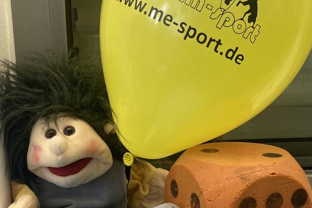 Plüschfigur mit frecher Frisur sitzt neben orangenem Würfel und blauen sowie gelben Luftballons auf einer Fensterbank.