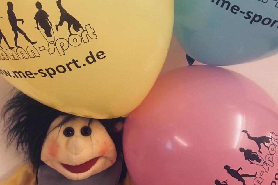 Stoffpuppe sitzt auf einer orangefarbenen Fl&auml;che, umgeben von gelben, blauen und rosa Luftballons mit Werbeaufdruck.