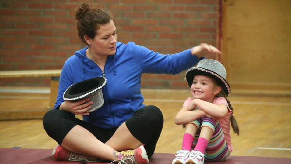 Frau mit einem schwarzen Hut sitzt auf einer Matte neben einem Mädchen mit einem glitzernden Hut, beide lächeln.