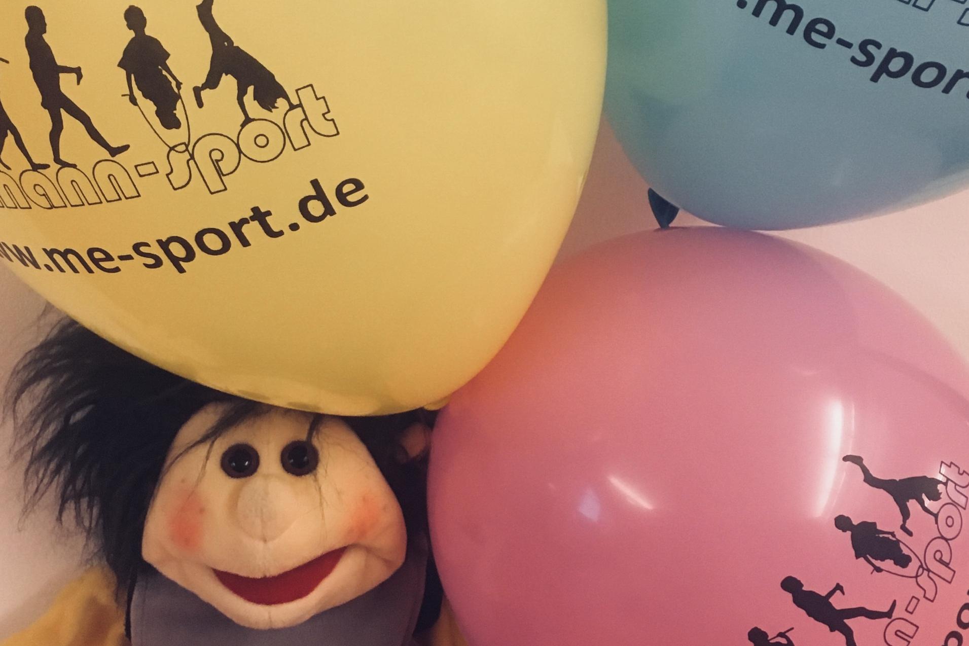 Stoffpuppe sitzt auf einer orangefarbenen Fläche, umgeben von gelben, blauen und rosa Luftballons mit Werbeaufdruck.