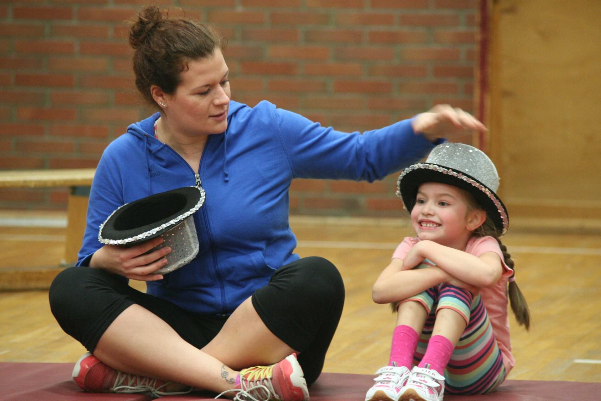 Frau mit einem schwarzen Hut sitzt auf einer Matte neben einem M&auml;dchen mit einem glitzernden Hut, beide l&auml;cheln.