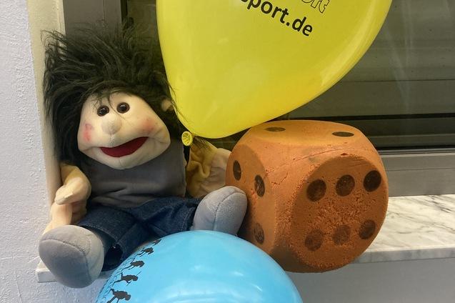 Plüschfigur mit frecher Frisur sitzt neben orangenem Würfel und blauen sowie gelben Luftballons auf einer Fensterbank.