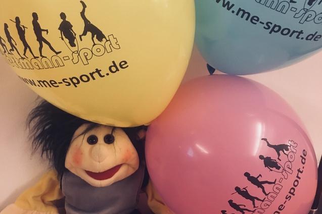 Stoffpuppe sitzt auf einer orangefarbenen Fläche, umgeben von gelben, blauen und rosa Luftballons mit Werbeaufdruck.