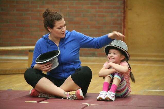 Frau mit einem schwarzen Hut sitzt auf einer Matte neben einem Mädchen mit einem glitzernden Hut, beide lächeln.