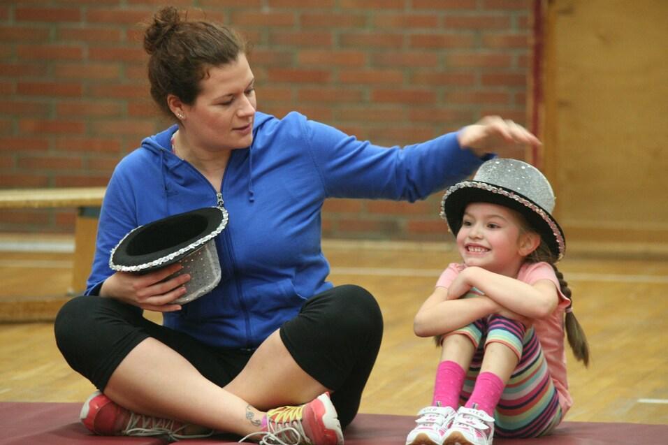 Frau mit einem schwarzen Hut sitzt auf einer Matte neben einem M&auml;dchen mit einem glitzernden Hut, beide l&auml;cheln.