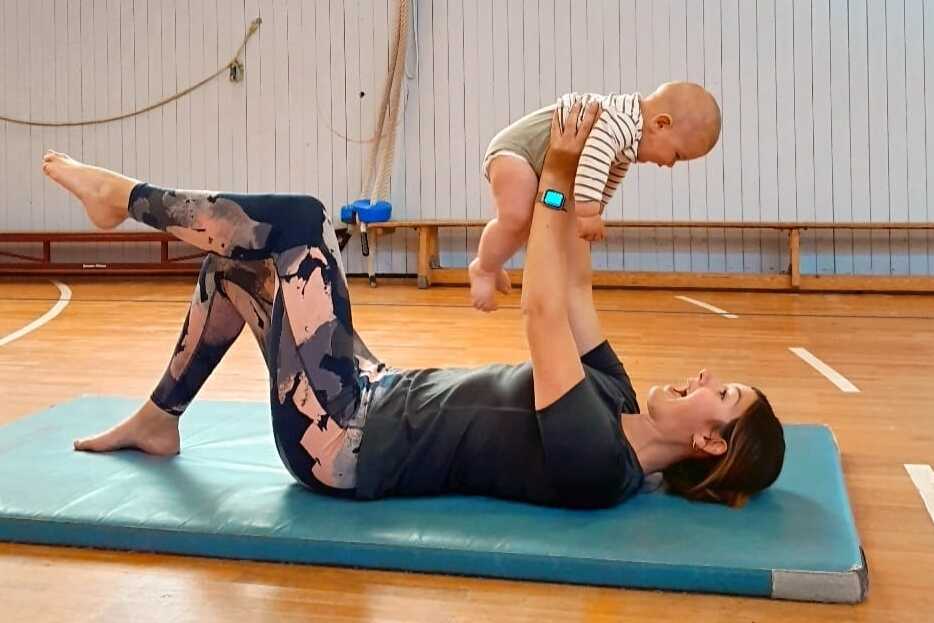Mutter liegt auf einer Matte und hebt spielend ihr Baby über ihr Gesicht in einer Turnhalle.