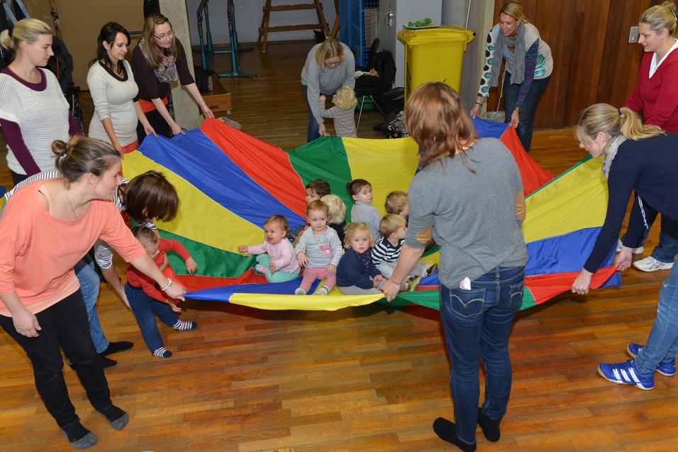 Gruppevon Erwachsenen und Kleinkindern, die gemeinsam mit einem bunten Parachute spielen und auf einem Holzfußboden sitzen.