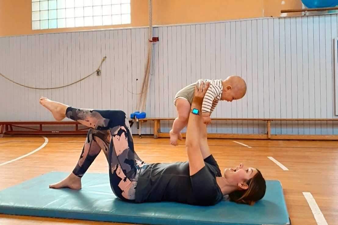 Mutter liegt auf einer Matte und hebt spielend ihr Baby &uuml;ber ihr Gesicht in einer Turnhalle.