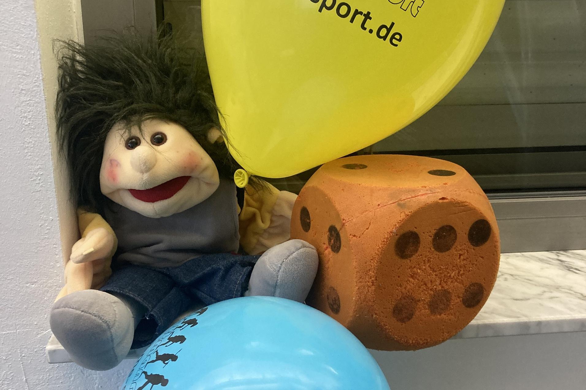 Plüschfigur mit frecher Frisur sitzt neben orangenem Würfel und blauen sowie gelben Luftballons auf einer Fensterbank.