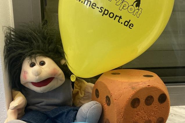 Plüschfigur mit frecher Frisur sitzt neben orangenem Würfel und blauen sowie gelben Luftballons auf einer Fensterbank.