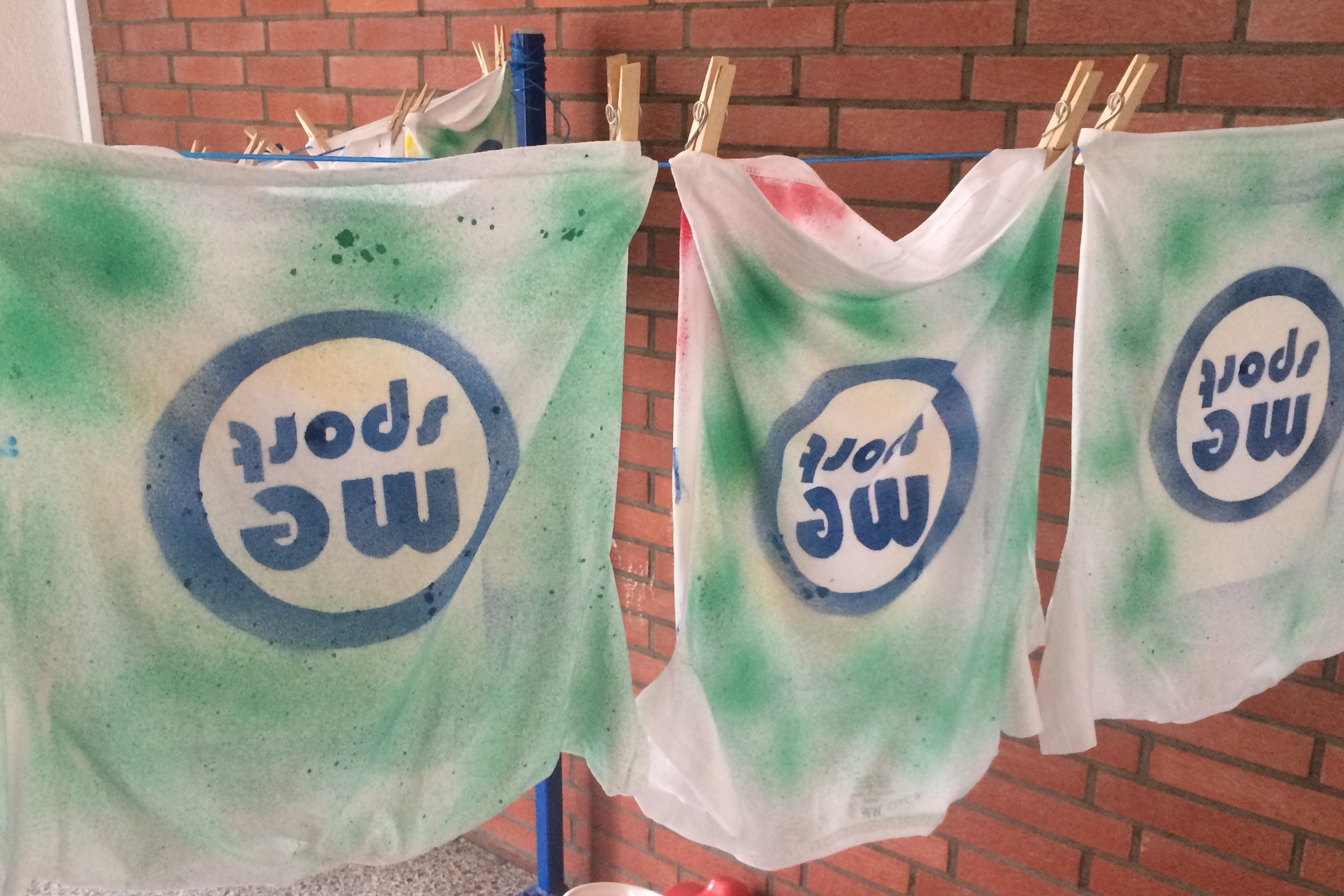 Drei weiße T-Shirts mit grünem und buntem Spraydruck hängen an einer Wäscheleine neben einer roten Spielzeugautobank.