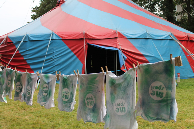 Bunte Shirts mit dem Aufdruck "mc sport" hängen an einer Leine vor einem rot-blauen Zelt.