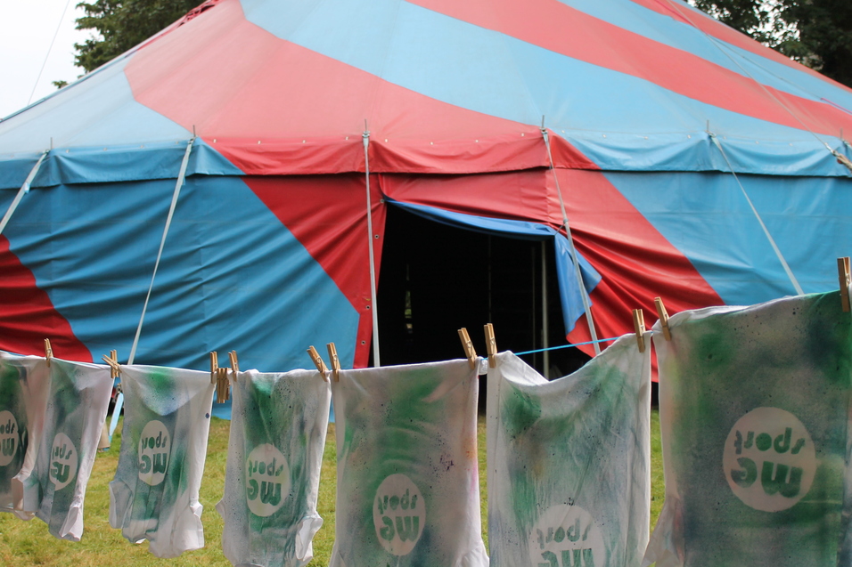 Bunte Shirts mit dem Aufdruck "mc sport" hängen an einer Leine vor einem rot-blauen Zelt.