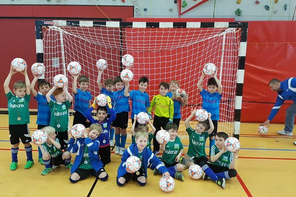 Gruppe von Kindern in Sportkleidung hält Fußball über Kopf vor einem Tor in einer Sporthalle.