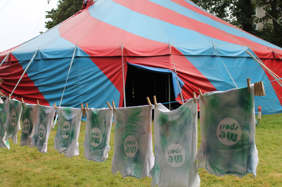 Bunte Shirts mit dem Aufdruck "mc sport" hängen an einer Leine vor einem rot-blauen Zelt.