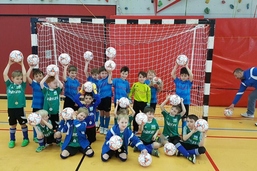 Gruppe von Kindern in Sportkleidung hält Fußball über Kopf vor einem Tor in einer Sporthalle.