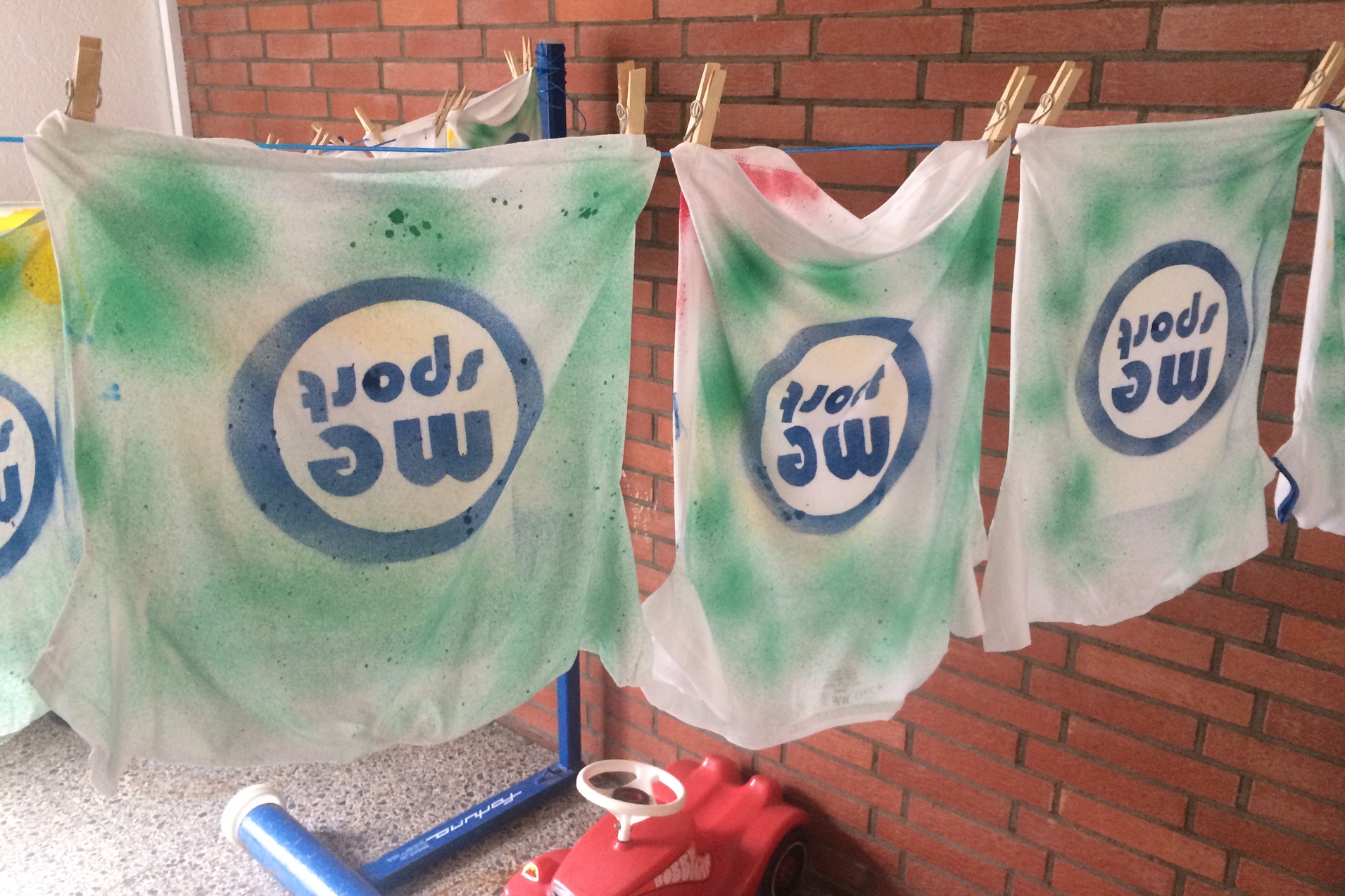 Drei weiße T-Shirts mit grünem und buntem Spraydruck hängen an einer Wäscheleine neben einer roten Spielzeugautobank.