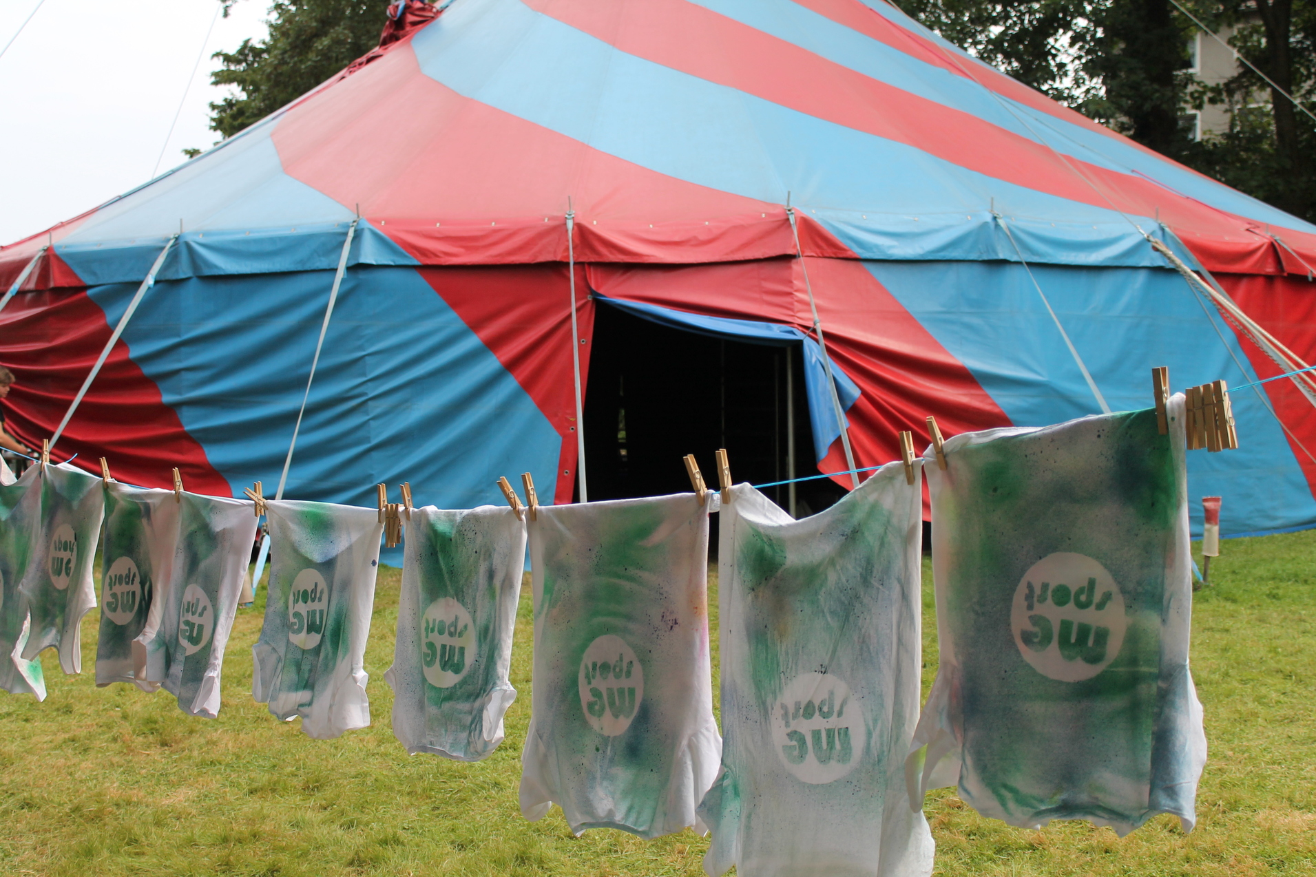 Bunte Shirts mit dem Aufdruck "mc sport" hängen an einer Leine vor einem rot-blauen Zelt.
