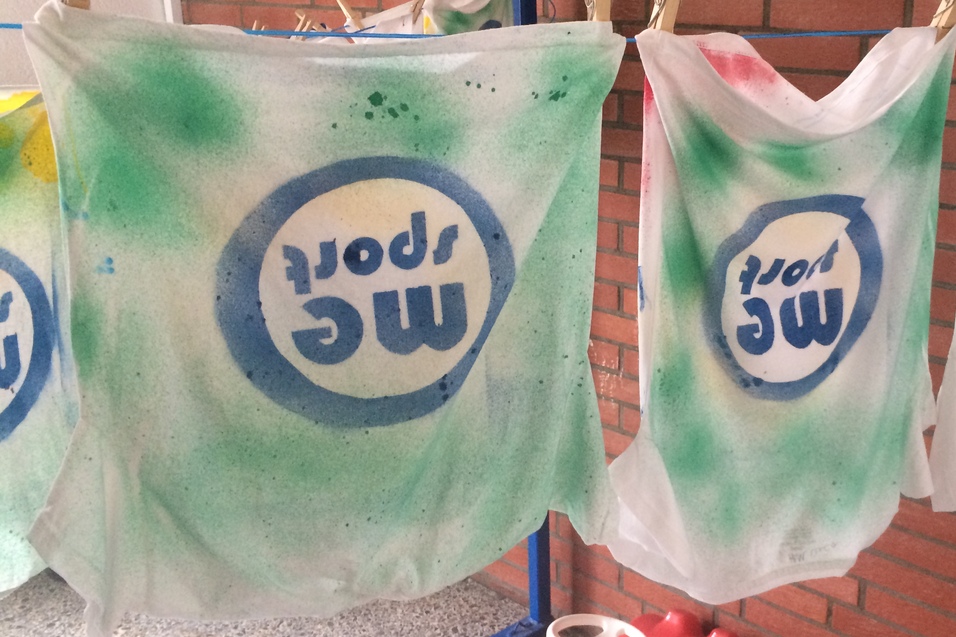 Drei weiße T-Shirts mit grünem und buntem Spraydruck hängen an einer Wäscheleine neben einer roten Spielzeugautobank.