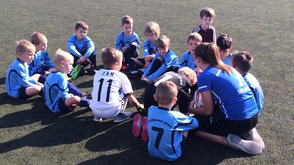 Gruppe von Kindern mit Trainingsanzügen sitzt auf einem Fußballplatz und hört einem Trainer zu.