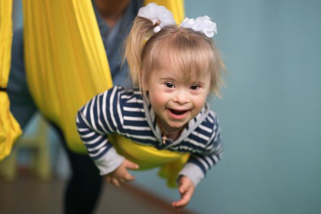 Lächelndes Kind mit blonden Haaren in einer gelben Hängematte, trägt einen gestreiften Pullover und eine weiße Haarschleife.