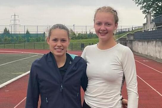 Zwei Läuferinnen stehen auf einer roten Laufbahn, sportlich gekleidet, lächeln und posieren nebeneinander.