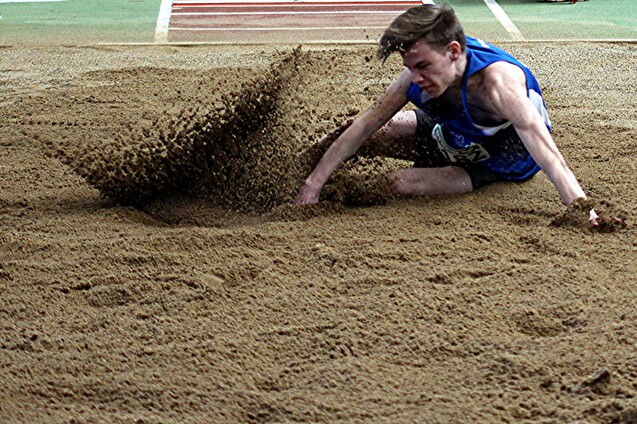Springer landet in einem Sandgrube, während Sandpartikel aufsteigen; er trägt ein blaues Trikot und ist im Wettkampf.