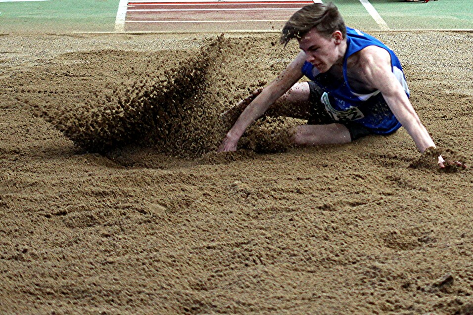 Springer landet in einem Sandgrube, während Sandpartikel aufsteigen; er trägt ein blaues Trikot und ist im Wettkampf.