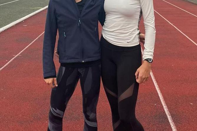 Zwei Läuferinnen stehen auf einer roten Laufbahn, sportlich gekleidet, lächeln und posieren nebeneinander.