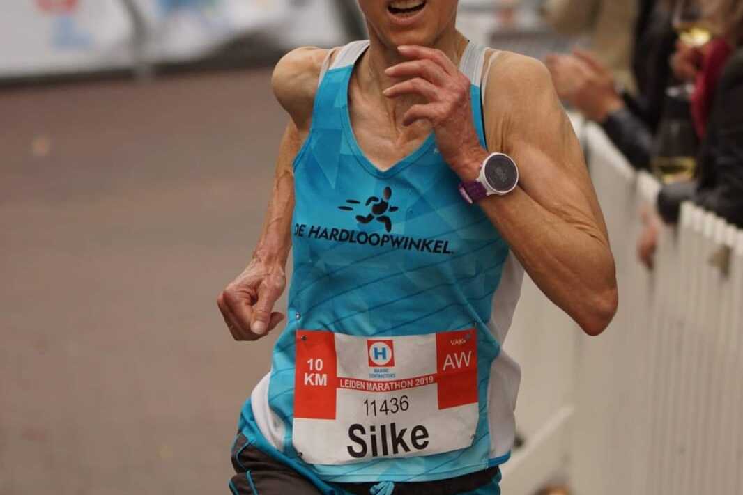 Läuferin im blauen Shirt mit Sonnenbrille, die beim Leiden Marathon den 10-km-Lauf absolviert. Zuschauer applaudieren im Hintergrund.