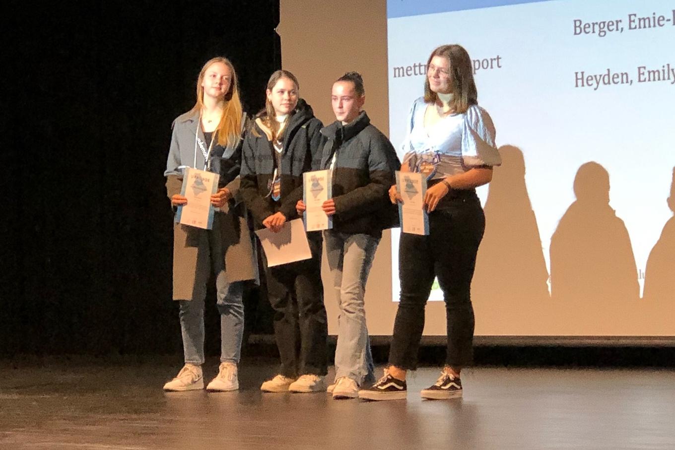 Vier junge Frauen stehen auf einer Bühne und halten Urkunden während einer Preisverleihung.