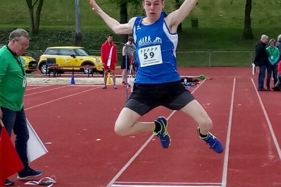 Junger Athlet springt in eine Sandgrube beim Weitsprung-Wettbewerb auf einem Sportplatz. Zuschauer im Hintergrund.