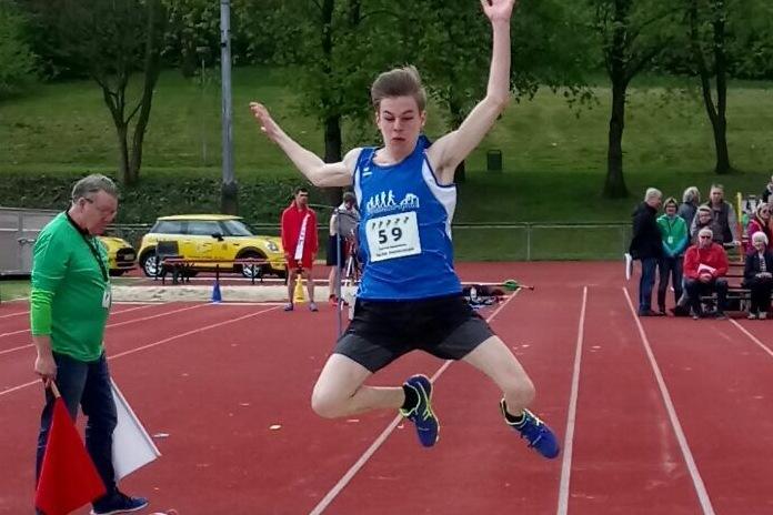 Junger Athlet springt in eine Sandgrube beim Weitsprung-Wettbewerb auf einem Sportplatz. Zuschauer im Hintergrund.