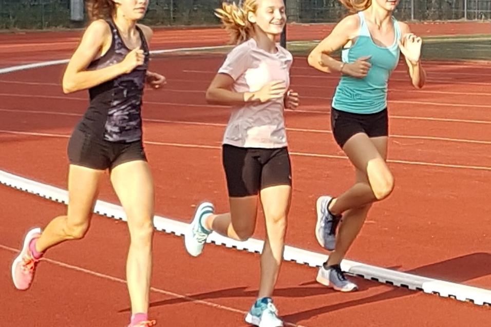 Drei Athletinnen laufen auf einer Laufbahn, während die Sonne auf die rote Bahn scheint.