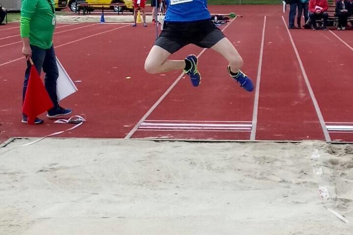Junger Athlet springt in eine Sandgrube beim Weitsprung-Wettbewerb auf einem Sportplatz. Zuschauer im Hintergrund.