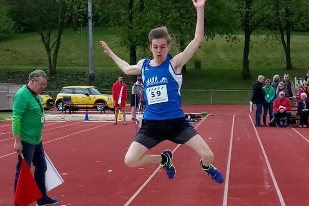 Junger Athlet springt in eine Sandgrube beim Weitsprung-Wettbewerb auf einem Sportplatz. Zuschauer im Hintergrund.
