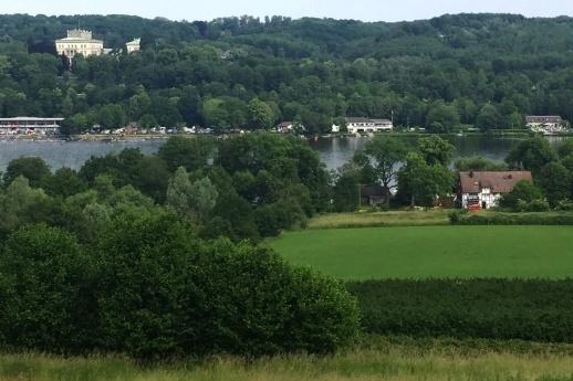 Panorama einer grünen Landschaft mit einem See, umgeben von Bäumen und mehreren Gebäuden im Hintergrund.