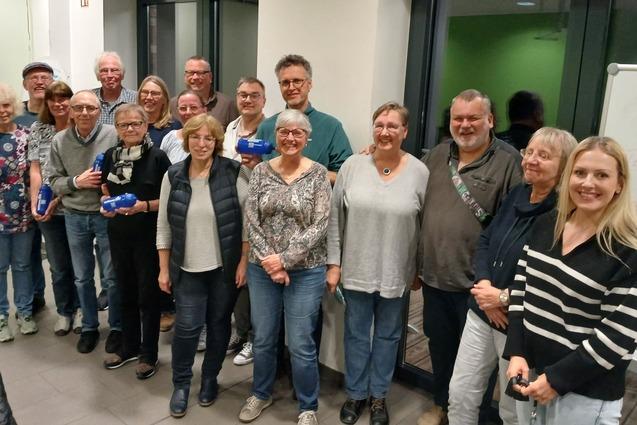 Gruppe von 20 Personen in einem Raum, lächelnd und stehend, mit blauen Objekten in den Händen, hinter ihnen Fenster und Tafel.