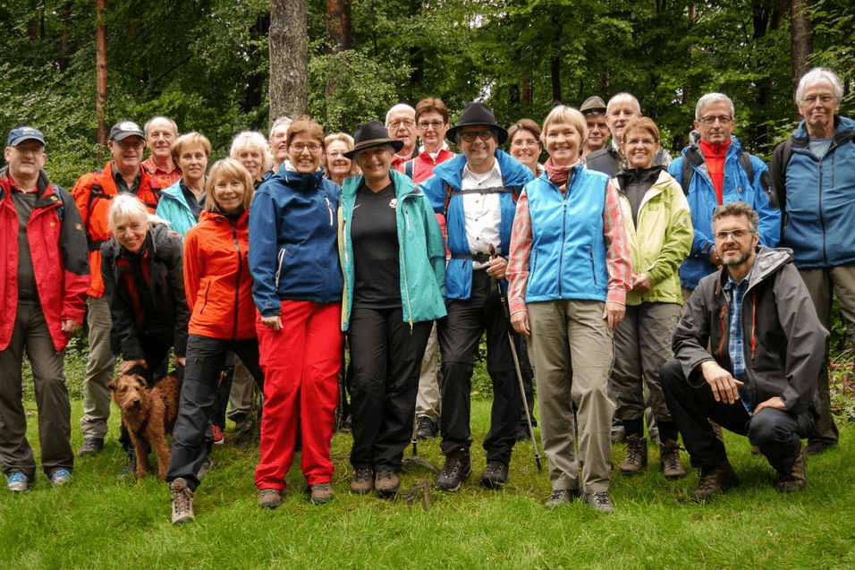 Gruppe von Wanderern mit unterschiedlichen Outfits steht auf einer grünen Wiese im Wald, begleitet von einem Hund.