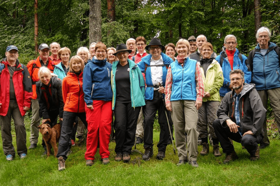 Gruppe von Wanderern mit unterschiedlichen Outfits steht auf einer grünen Wiese im Wald, begleitet von einem Hund.