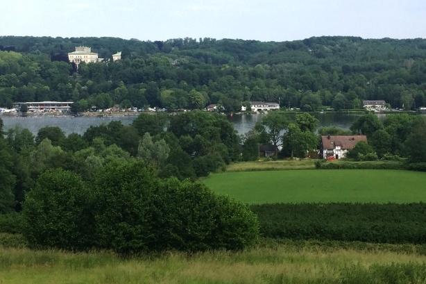 Panorama einer grünen Landschaft mit einem See, umgeben von Bäumen und mehreren Gebäuden im Hintergrund.