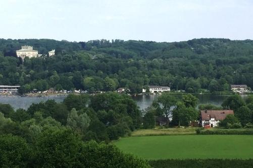 Panorama einer grünen Landschaft mit einem See, umgeben von Bäumen und mehreren Gebäuden im Hintergrund.