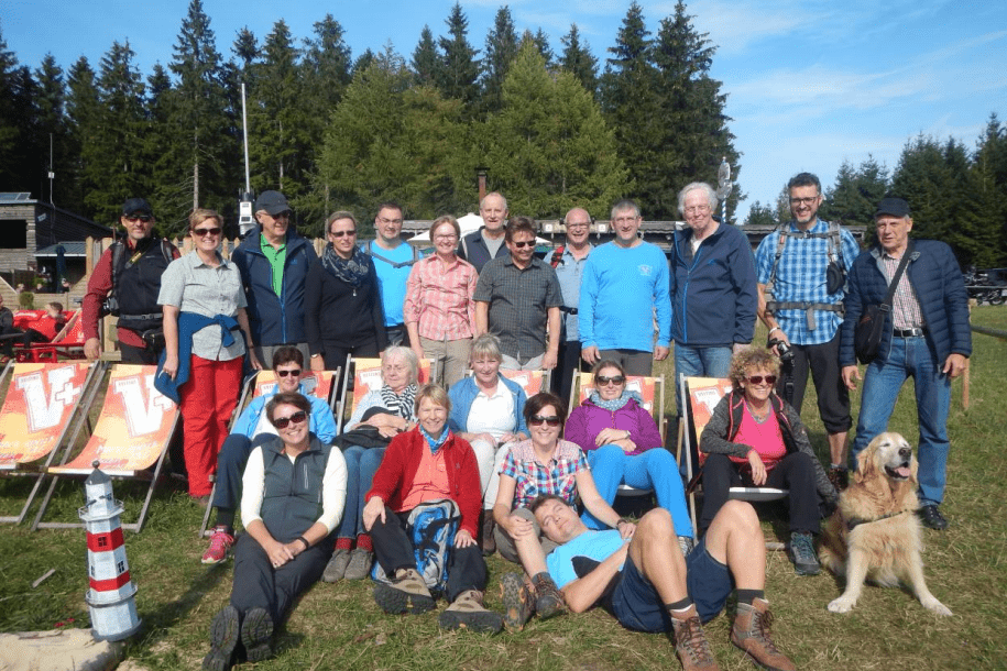 Gruppe von 20 Menschen und einem Hund im Freien, sitzend auf Liegestühlen vor Waldkulisse, sonniger Tag.