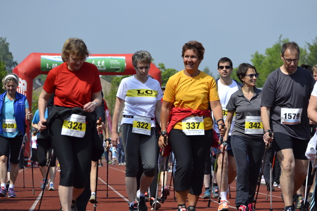 Gruppe von Menschen in Sportkleidung geht mit Nordic-Walking-St&ouml;cken auf einer Laufbahn, im Hintergrund ein Torbogen.