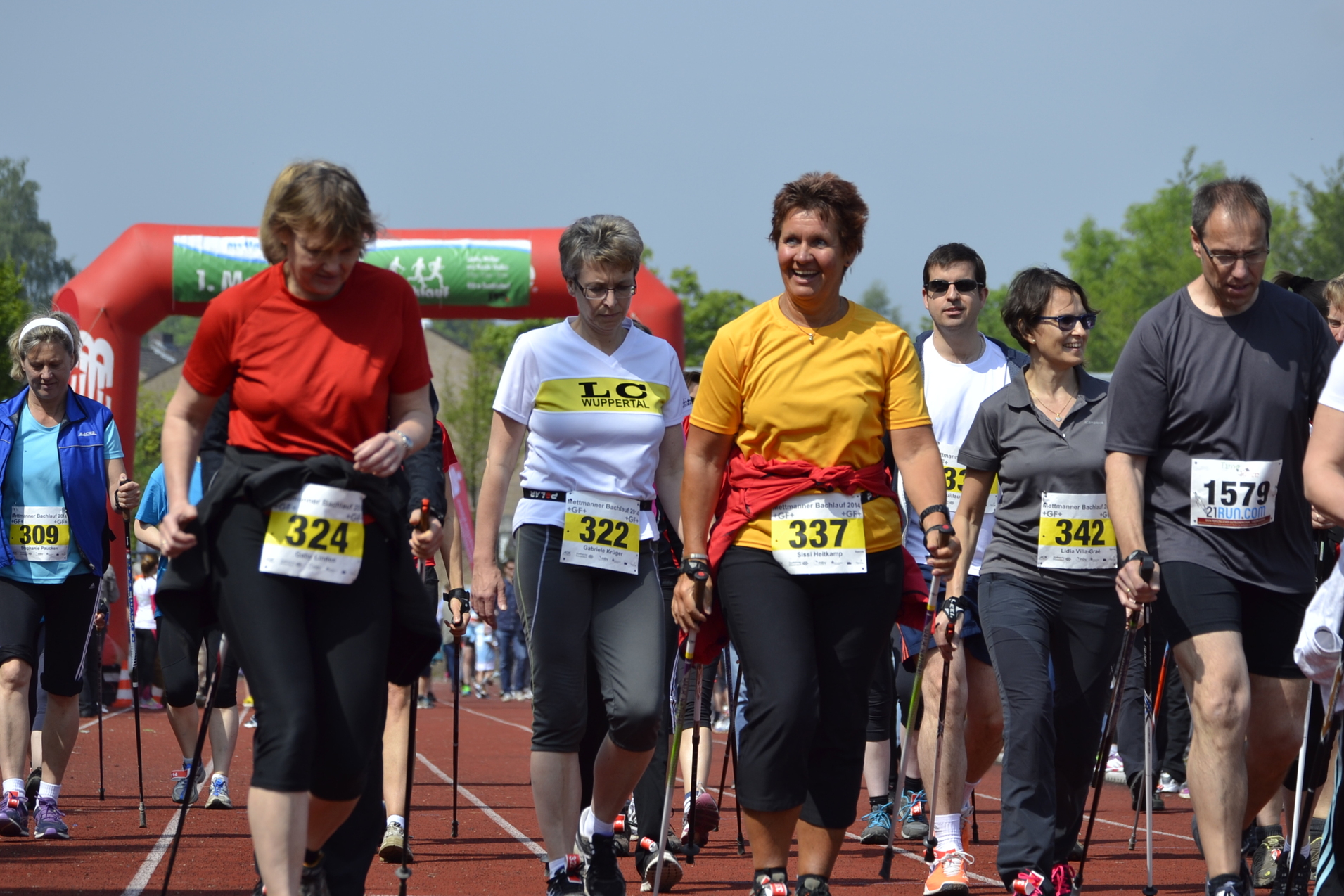 Gruppe von Menschen in Sportkleidung geht mit Nordic-Walking-St&ouml;cken auf einer Laufbahn, im Hintergrund ein Torbogen.