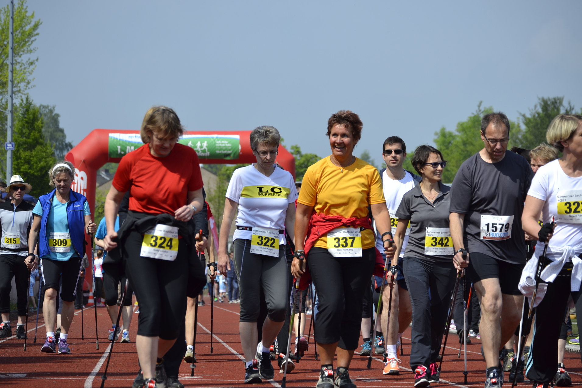 Gruppe von Menschen in Sportkleidung geht mit Nordic-Walking-Stöcken auf einer Laufbahn, im Hintergrund ein Torbogen.