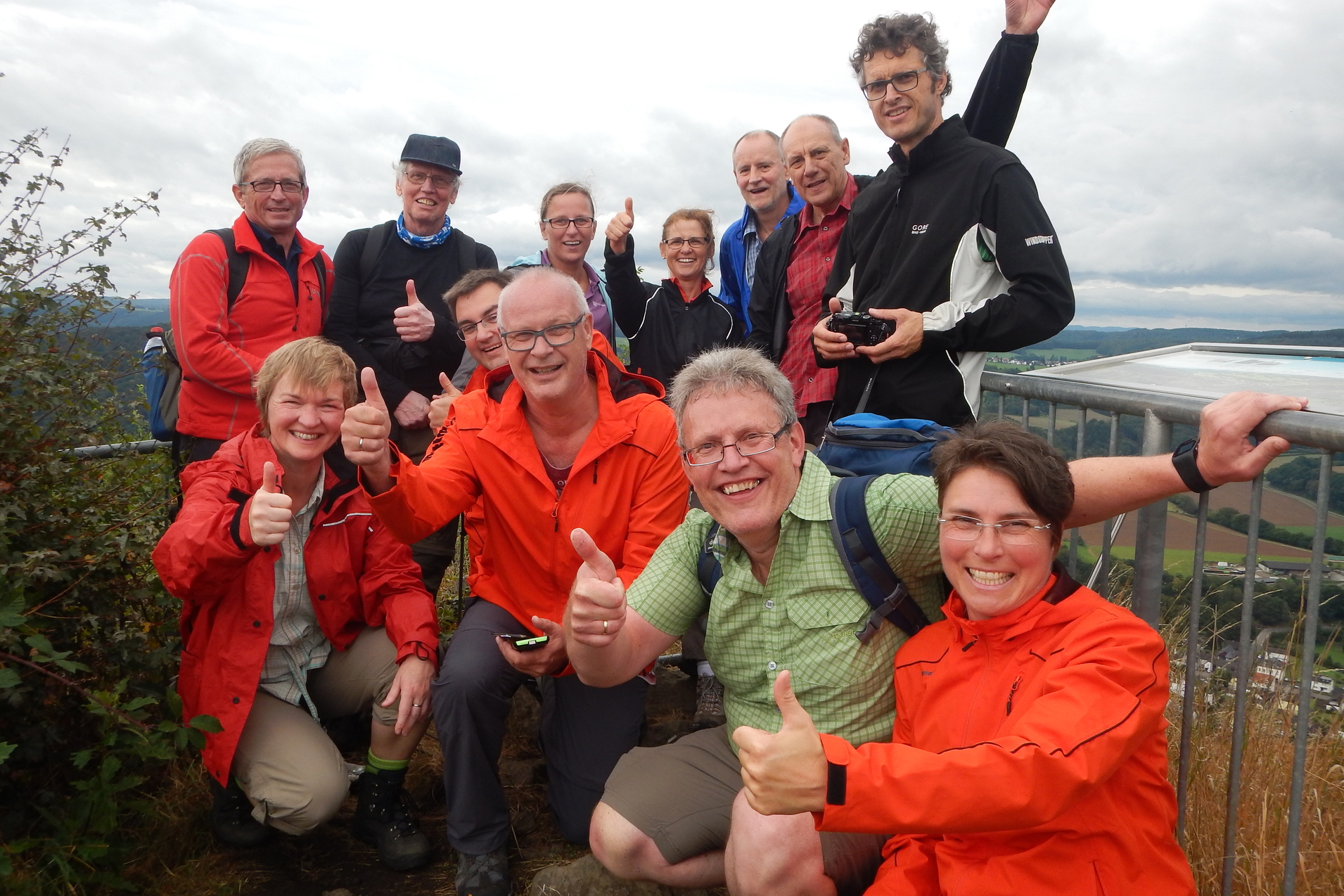 Gruppe von zehn Personen in Wanderkleidung, lächelnd und mit Daumen hoch, an einem Aussichtspunkt im Freien.