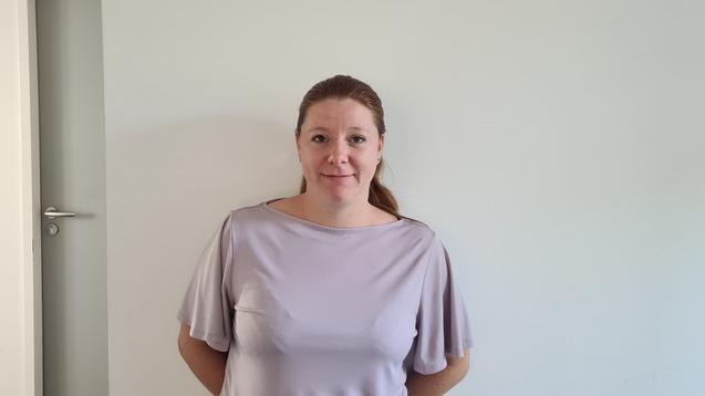 Frau mit langen Haaren steht lächelnd an einer Wand in einem hellen Raum, neben einer geschlossenen Tür und einem Feuerlöscher.